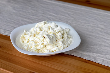 A white square plate filled with cottage cheese stands on a linen tablecloth on a wooden table. Selective focus