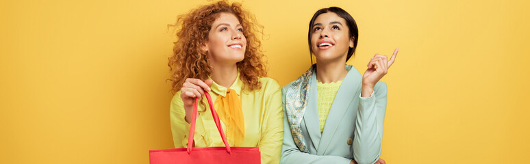 panoramic shot of happy redhead girl holding shopping bag near smiling african american woman pointing with finger isolated on yellow