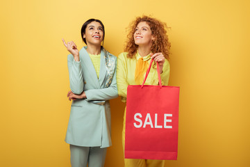 happy redhead girl holding shopping bag with sale lettering near smiling african american woman...