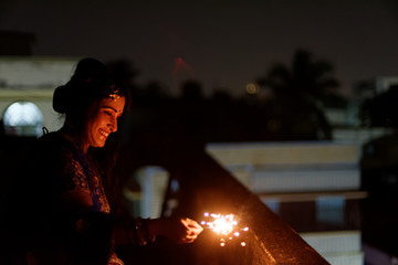 Young and beautiful Indian Bengali/gujarati/rajasthani woman in Indian traditional dress is celebrating Diwali with fire crackers on rooftop in darkness. Indian lifestyle and Diwali celebration