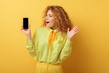 excited redhead woman looking at smartphone with blank screen on yellow