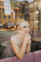 Beautiful pretty attractive inspired tender young sexy blonde girl in a light pink puffy dress looks out the window and looks at the street sitting in a pink cafe, portrait of the bride