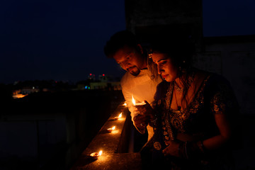 Young and beautiful Indian Gujarati couple in Indian traditional dress lightening Diwali diya/lamps sitting on the terrace in blue hour on Diwali evening. Indian lifestyle and Diwali celebration