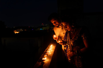 Young and beautiful Indian Gujarati couple in Indian traditional dress lightening Diwali diya/lamps sitting on the terrace in darkness on Diwali evening. Indian lifestyle and Diwali celebration