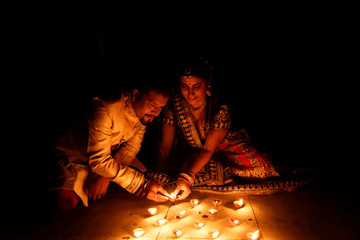 Beautiful Indian Gujarati couple in Indian traditional dress lightening Diwali diya/lamps sitting on the floor in darkness on Diwali evening. Indian lifestyle and Diwali celebration