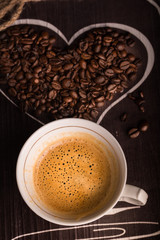 A cup of delicious hot coffee accompanied by a heart filled with coffee beans. Vertical format