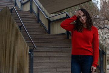 Portrait of worried attractive young woman keeps hand on forhead, in the park, orange sweater and jeans, long curly hair. Place for your text in copy space.