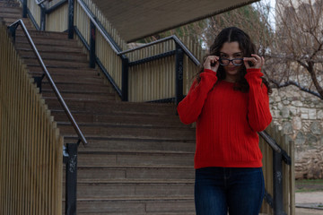 Portrait of pretty caucasian young woman taking off eyeglasses, holding frames near forehead as gazing at camera, in the park, orange sweater and jeans, long curly hair. Place for your text in copy sp