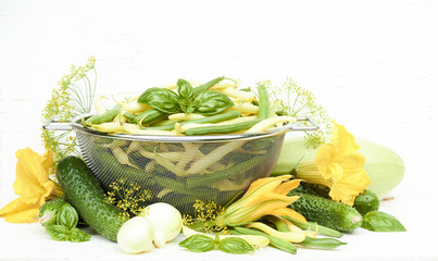 Variety of first green vegetables and herbs: cucumber, green string bean, basil, zucchini, dill, onion and yellow flowers isolated on white wooden background, closeup, copy space