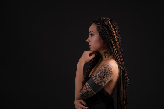 Shot of a young woman with a tattoo on her arms and with braids against  dark background with space in the studio