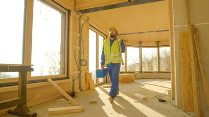 CLOSE UP: Contractor in blue overalls carries a toolbox and ladder across room