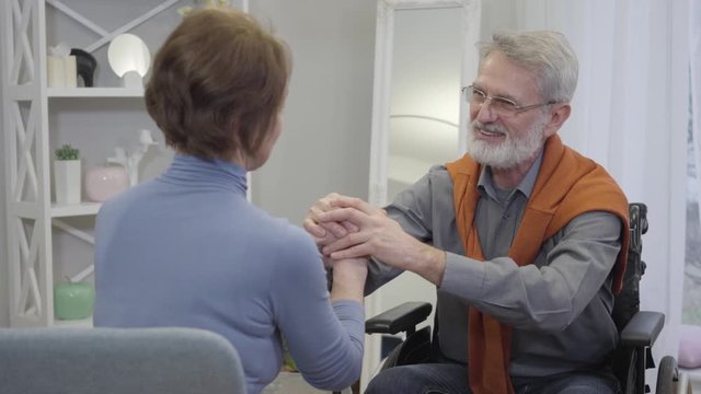 Elderly handsome Caucasian man holding woman's hands, talking and smiling. Happy senior retirees enjoying time together in nursing home.