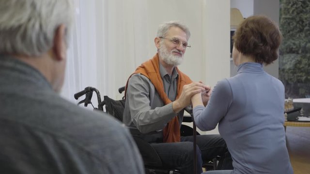 Graceful old Caucasian man in wheelchair holding hands of senior woman. Mature retiree in love flirting with female resident of nursing home. Lonely man reading at the foreground.