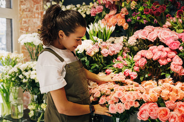 Woman florist wearing apron looking on flowers in her store - obrazy, fototapety, plakaty
