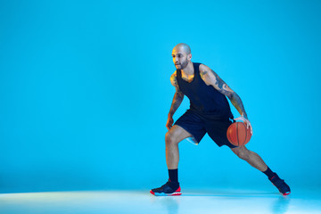 Young basketball player of team wearing sportwear training, practicing in action, motion isolated on blue background in neon light. Concept of sport, movement, energy and dynamic, healthy lifestyle.