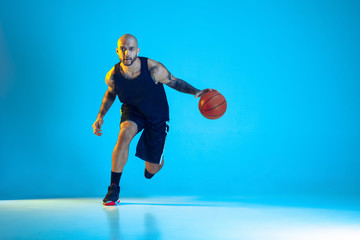 Fototapeta na wymiar Young basketball player of team wearing sportwear training, practicing in action, motion isolated on blue background in neon light. Concept of sport, movement, energy and dynamic, healthy lifestyle.