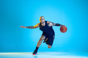 Fototapeta na wymiar Young basketball player of team wearing sportwear training, practicing in action, motion isolated on blue background in neon light. Concept of sport, movement, energy and dynamic, healthy lifestyle.