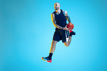 Fototapeta na wymiar In jump. Young basketball player of team wearing sportwear training, practicing in action, motion on blue background in neon light. Concept of sport, movement, energy and dynamic, healthy lifestyle.