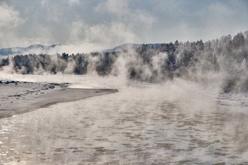 Winter landscape. Not freezing river with snowy trees along the banks. Steam rises from the winter mountain river against the blue sky. Morning back light. Russia. Altai Republic.