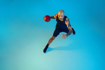 Fototapeta premium Top view. Young basketball player of team wearing sportwear training, practicing in action, motion on blue background in neon light. Concept of sport, movement, energy and dynamic, healthy lifestyle.
