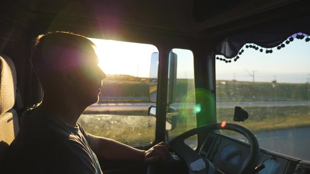 Man driving a truck and carefully watching the road. Caucasian guy is riding through the countryside on the sunset background. Profile of lorry driver inside the cab. Side view Slow motion Close up