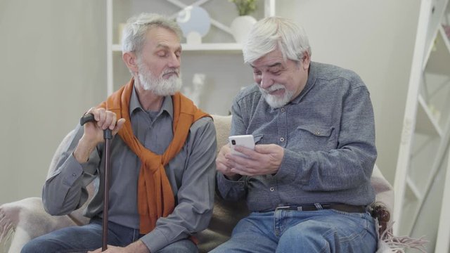 Active old Caucasian man showing photos on smartphone to friend with walking stick. Positive male retirees spending time together in nursing home.