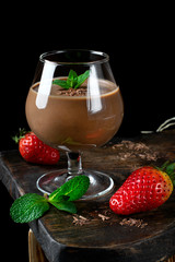 Chocolate pudding topped with mint in the glass against the black background. Ripe strawberry on the wooden board
