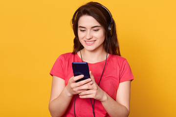 Indoor shot of young girl listening and enjoying music, holding cellular phone in hands and earphones on top of her head, girl wearing red casual t shirt, model posing isolated over yellow background.