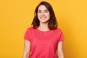 Image of attractive woman with dark hair being very glad, looking with broad smile showing her perfect teeth, excited female wearing red casual t shirt, posing isolated over yellow background.