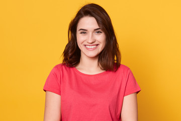 Fototapeta premium Picture of Caucasian brunette woman posing isolated over yellow background, dark haired lady looks smiling directly at camera, looking happy, lady wearing red t shirt. People emotions concept.