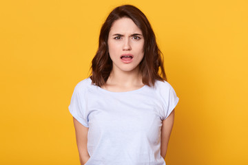 Horizontal shot of young beautiful woman wearing white casual t shirt standing isolated over yellow background, looks scared, having surprise face, afraid of something, caucasian female has dark hair.