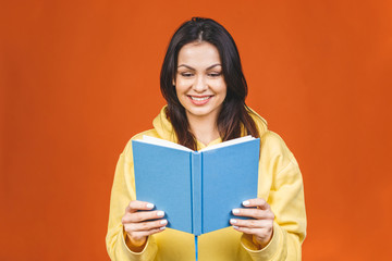 Beautiful young woman wearing casual standing isolated over orange background, reading a book.
