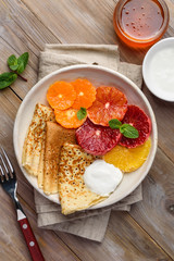 Crepes  with citrus, sour cream and honey for breakfast. Thin pancakes with slices of orange and mandarin on a plate on a wooden table. Top view