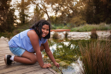 A 9 year old girl wearing jean shorts who is leaning at the edge of a wooden deck next to a pond, during a sunset.