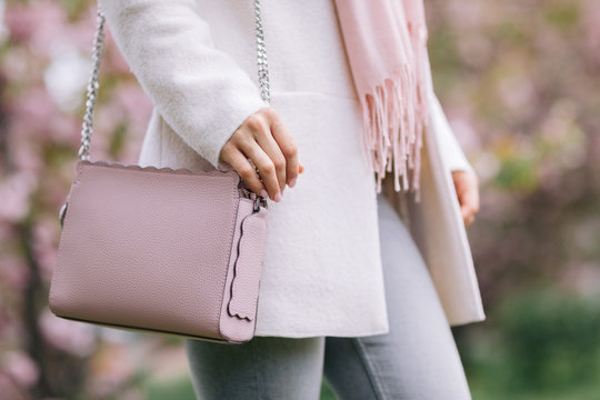 woman in white coat, scarf and pink handbag posing in the park with blooming spring trees.