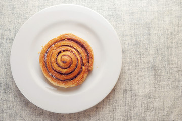 One tasty cinnamon roll on a white plate. Bakery product on a simple table cloth. Top view.