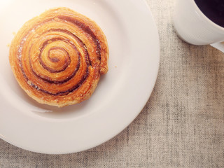 One crispy cinnamon roll on a white plate and a white cup with coffee. Concept breakfast.