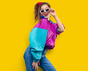 Cool teenager. Fashionable DJ girl in colorful trendy jacket and vintage retro sunglasses enjoys style of 80s � 90s vibes. Teenager Girl at disco party. Young fashion model on yellow color background. - obrazy, fototapety, plakaty