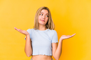 Young blonde caucasian woman isolated on a yellow background doubting and shrugging shoulders in questioning gesture.