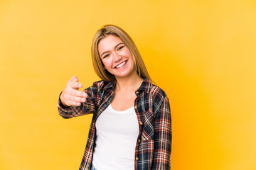 Young blonde caucasian woman isolated on a yellow background stretching hand at camera in greeting gesture.