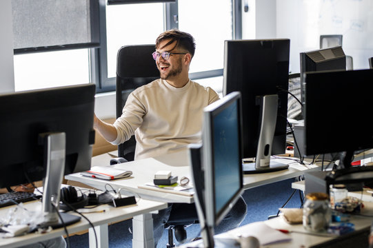 Young  Programmer Sitting At The Desk In His Office And Working .He Talk With Someone And Smiling.