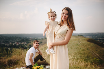 Family, mom, dad, daughter on picnic on a hill in the field. Mom and daughter in yellow identical dresses. Sunflowers.