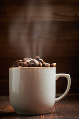 A white cup with coffee beans and steam over it on a wooden background.