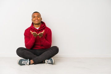 Young latin man sitting on the floor isolated holding something with palms, offering to camera.