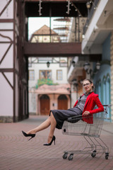 Beautiful cute young businesswoman in red jacket and formal suit and high-heeled shoes and glasses sits in shopping cart on blurred background of buildings