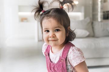 One year old baby is looking at the camera. Baby with two ponytails in a pink sundress. Cute girl sitting on a sofa