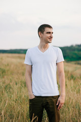 A guy in a white t-shirt and brown trousers in a field on a hill. Man portrait in nature. Short haircut, brunette.