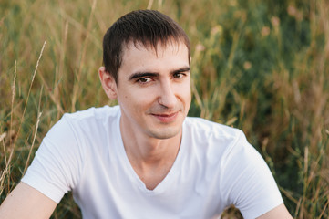 Naklejka na ściany i meble A guy in a white t-shirt and brown trousers in a field on a hill. Man portrait in nature. Short haircut, brunette.