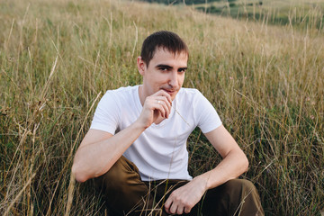 Naklejka na ściany i meble A guy in a white t-shirt and brown trousers in a field on a hill. Man portrait in nature. Short haircut, brunette.