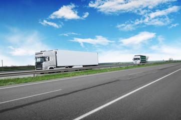 A big white truck and trailer with other truck on the countryside road against a blue sky with...